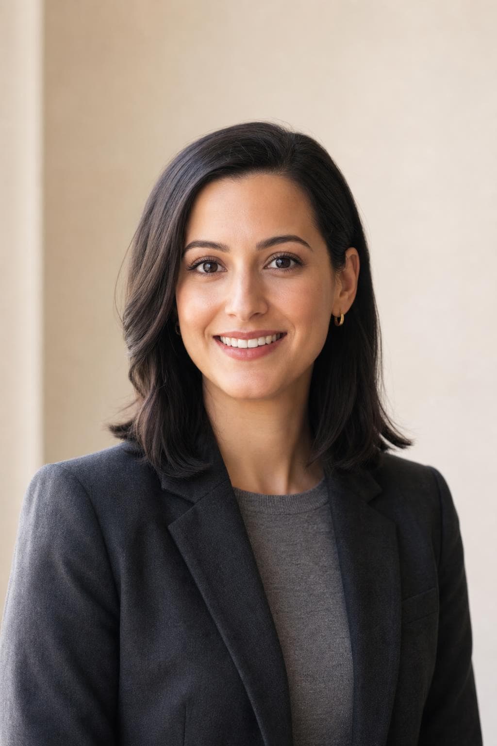 AI profile picture in LinkedIn style — woman in a charcoal blazer over a grey knit, bright neutral wall, even frontal light